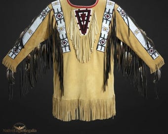 Puede incluir: Una camisa de manga larga color canela con largos flecos negros a lo largo de las mangas y el dobladillo inferior. La prenda incluye paneles de cuentas blancas en las mangas y un panel triangular en el pecho con patrones geométricos de cuentas.