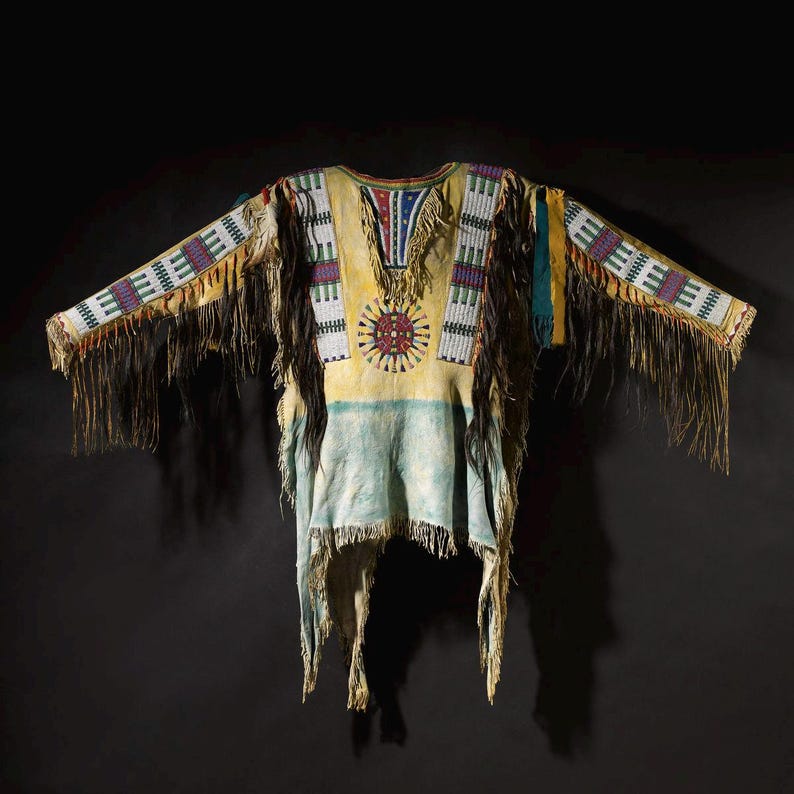 May include: A Native American-style tunic with long sleeves and fringed edges. The garment features a yellow body with a sunburst design, beaded accents, and colorful geometric patterns. The sleeves are adorned with white and purple beadwork.