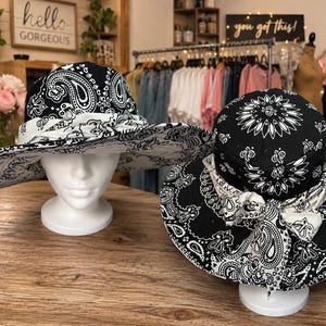Könnte beinhalten: Zwei breitkrempige Hüte mit schwarz-weißem Paisley-Bandana-Muster. Jeder Hut ist mit einem weißen Stoffband verziert, das vorne zu einer Schleife gebunden ist. Ein Hut wird auf einem weißen Schaufensterpuppenkopf präsentiert, der andere steht auf einem weißen Ständer.