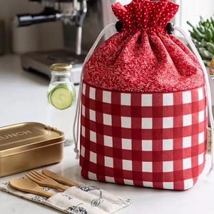 May include: A cylindrical drawstring lunch bag featuring a red and white checkered pattern on the lower half and a red fabric with a white floral and polka dot print on the upper section, secured with white cords and black toggles.