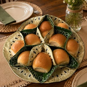 May include: A fabric bread warmer with a star-shaped design featuring a light olive green and dark green paisley bandana print. The warmer contains eight golden-brown dinner rolls nestled within its individual triangular pockets.