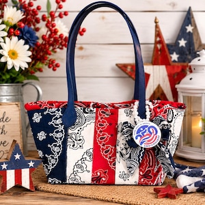 May include: A quilted tote bag featuring a red, white, and blue bandana print pattern with vertical stripes. The bag has two dark blue handles and a decorative bow attached to one handle, which includes a round pin reading 25th Anniversary.