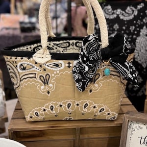 May include: A quilted fabric tote bag featuring a tan and black paisley bandana print. The bag has two thick, cream-colored rope handles and a black fabric trim. A black paisley bow is attached to one handle with a small turquoise stone charm.