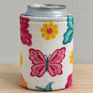 Peut inclure: Une housse isotherme en tissu blanc pour canette, ornée d'un motif répétitif de papillons roses, de fleurs jaunes et d'ailes de papillon bleu sarcelle. La housse est cylindrique et s'ajuste autour d'une canette en métal argenté.