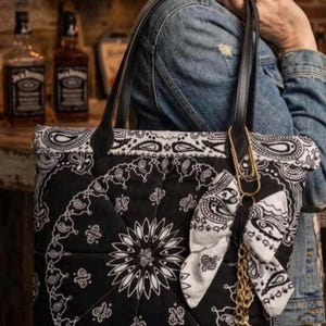 May include: A black and white quilted tote bag featuring a classic bandana paisley print. The bag has two black shoulder straps, a decorative white bandana fabric bow attached to a gold-toned paperclip charm, and a dangling gold-toned boot charm.