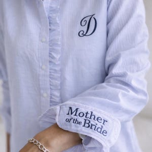 May include: Light blue striped button-down shirt with a ruffled collar and cuffs. The cuff is embroidered with "Mother of the Bride" in navy blue. A navy blue letter "D" is embroidered on the chest.