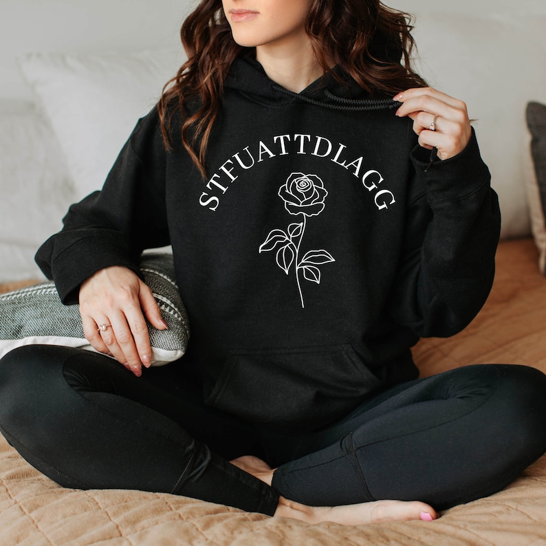 K&ouml;nnte beinhalten: Schwarzer Kapuzenpullover mit einer wei&szlig;en Rose und dem Text "STFUATTDLAGG" auf der Vorderseite.