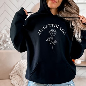 K&ouml;nnte beinhalten: Schwarzer Hoodie mit einer wei&szlig;en Rose und dem Text "STFUATTDLAGG" auf der Vorderseite.