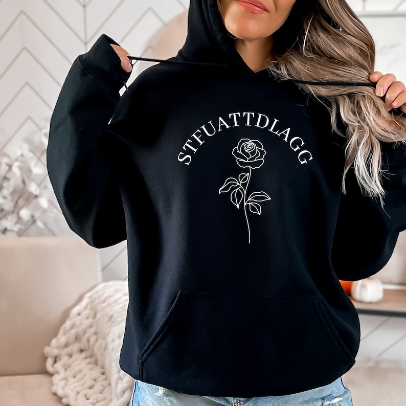 K&ouml;nnte beinhalten: Schwarzer Hoodie mit einer wei&szlig;en Rose und dem Text "STFUATTDLAGG" auf der Vorderseite.