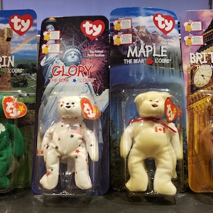 May include: Four Ty Beanie Babies, each in a plastic display case. The bears are Erin, Glory, Maple, and Britannia. Each bear is wearing a different outfit and has a different background image. The bears are all in the same pose, standing with their arms at their sides.