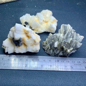 May include: Three raw mineral specimens. Two are white with black inclusions and yellow-brown hues. The third is a cluster of gray-green crystals. A stainless steel ruler is included for scale, showing measurements in inches.