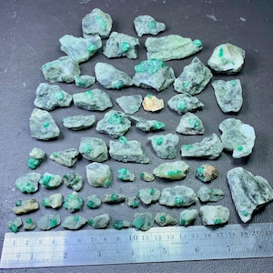 May include: A collection of rough, uncut emeralds embedded in matrix rock. The emeralds are a vibrant green, and the matrix is a mix of gray and white. A stainless steel ruler is placed next to the stones for scale.