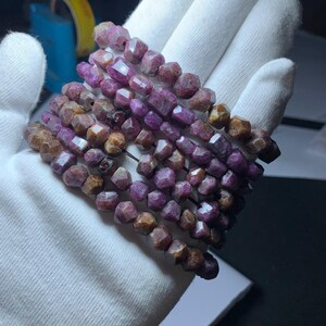 May include: A strand of pink and brown gemstone beads, likely ruby, strung on a wire. The beads are faceted and have a slightly irregular shape. The beads are arranged in a loose coil.