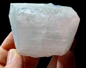 May include: A light pink and white crystal formation with a layered appearance. The crystal is held in a hand against a black background. The crystal has a rectangular shape with angled edges.