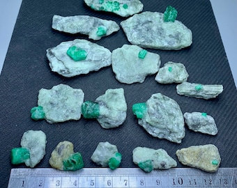 Op de afbeelding: Een verzameling ruwe smaragdmonsters, met levendige groene kristallen ingebed in lichtgekleurde rotsformaties. De smaragden variëren in grootte en vorm, met een liniaal voor schaal in centimeters. De rotsen zijn gerangschikt op een donkere ondergrond.