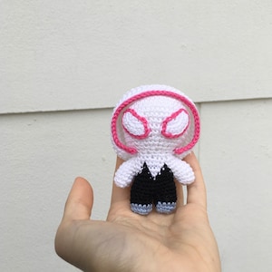 May include: A handmade crochet doll of a character with a white head and body, black shorts, and blue shoes. The doll has a pink outline around the eyes and head. The doll is held in a hand.
