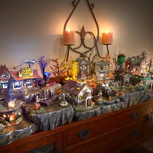 May include: A Halloween village display with miniature houses, trees, and figures. The village is set on a wooden table with a dark brown finish. The houses are lit with small lights and the scene is decorated with spooky details like a witch's hat and a sign that reads "Dead End Motel."