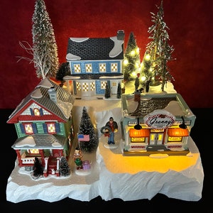 May include: A miniature Christmas village scene with a green house, a blue house, a store called "Granny's Jellies & Jams Est. 1900", and a lit Christmas tree. The scene is set on a snowy base with a white, rocky cliff.