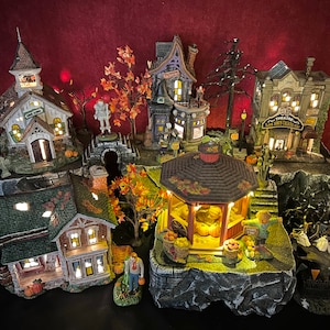 May include: A miniature Halloween village with a schoolhouse, a haunted house, a bakery, a gazebo, and a house with pumpkins. The village is lit up with lights and has a spooky autumn theme.