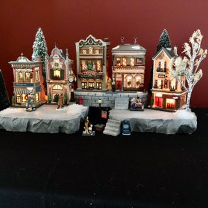 May include: A miniature village scene with a variety of buildings, including a theater, a store, and a church. The buildings are decorated with lights and snow, and there are small figures of people walking around. The scene is set on a rocky base with a small bridge and a river.