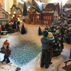 Pode incluir: Uma cena de vila de inverno em miniatura com um lago congelado, árvores cobertas de neve e pequenas casas. Há várias figuras de pessoas na cena, incluindo um casal de pé perto de uma árvore, uma criança patinando no gelo e um grupo de pessoas reunidas em torno de uma fogueira.