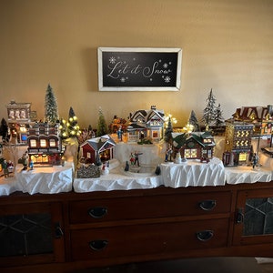 May include: A miniature Christmas village display with snow-covered buildings, trees, and a skating rink. The village is set on a wooden table with a sign that reads "Let it Snow".
