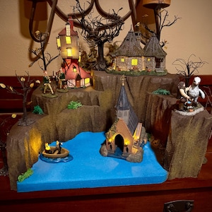 May include: A miniature Halloween village scene with illuminated buildings, including a church, houses, and a tower. The scene features a blue lake, figures, and bare trees, all set on a brown, rocky base.