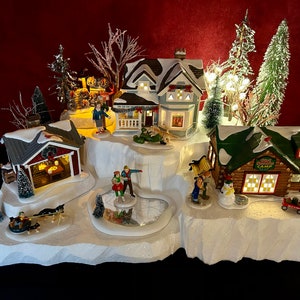 May include: A miniature winter village scene with a white, snowy base. The village features a blue house with a porch, a red cabin with a wreath, a log cabin with a green roof and the text "Ski Lodge Camp", and a frozen pond with two figures ice skating. The scene is lit with small white lights.