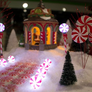Könnte beinhalten: Eine Miniatur-Winterdorfszene mit einem roten und weißen Zuckerstangen-Pfad, der zu einem kleinen Lebkuchenhaus führt. Die Szene ist mit weißem Schnee und kleinen immergrünen Bäumen dekoriert.