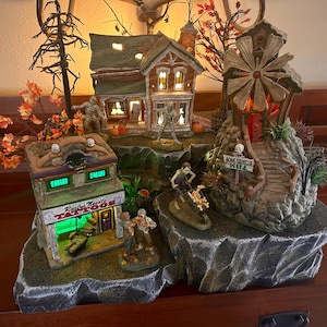 May include: A Halloween village scene with a haunted house, a tattoo parlor, a bone grinder mill, and a windmill. The scene is lit with green and orange lights.