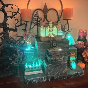 May include: A Halloween-themed display featuring a miniature haunted house, spooky trees, and glowing lights. The scene includes a miniature graveyard, pumpkins, and candle holders, creating a festive and eerie atmosphere. The color palette includes shades of green, orange, and black.