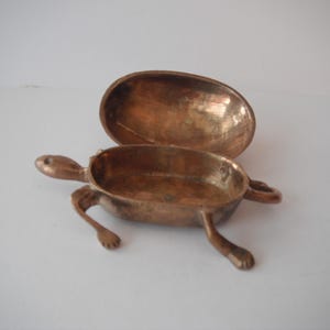 May include: A metallic, copper-toned turtle-shaped trinket box with an open, hinged lid. The object features a smooth, oval-shaped body, four distinct legs, and a small head extending from the front. The interior is hollow and metallic.