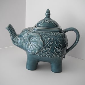 May include: A teal blue ceramic teapot shaped like an elephant with intricate floral designs. The teapot has a lid with a finial on top.