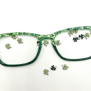 Op de afbeelding: Groene bril met een glitterend, gespikkeld ontwerp. De monturen zijn versierd met kleine, zilverkleurige klaverbedels. De lenzen zijn helder en de algehele esthetiek is feestelijk en decoratief.