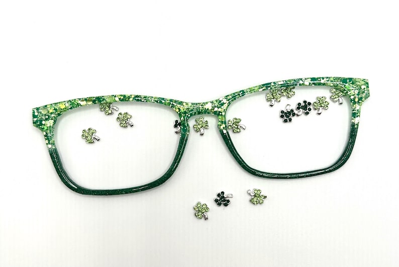 Op de afbeelding: Groene bril met een glitterend, gespikkeld ontwerp. De monturen zijn versierd met kleine, zilverkleurige klaverbedels. De lenzen zijn helder en de algehele esthetiek is feestelijk en decoratief.
