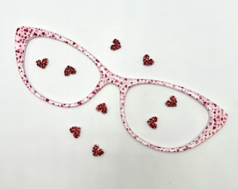 May include: Pink cat-eye glasses with a pattern of small red hearts. Scattered around the glasses are several small, glittery red heart-shaped embellishments. The glasses and hearts are set against a white background.