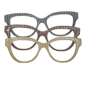 May include: Three pairs of eyeglasses with a retro cat-eye shape. The frames feature a gingham pattern in blue, brown, and tan. The glasses are arranged in a stacked formation, showcasing their unique design and style.