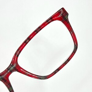 May include: Close-up of eyeglasses with a red plaid pattern. The frames are a vibrant red with black and green accents, creating a classic and stylish look. The glasses have a rectangular shape.