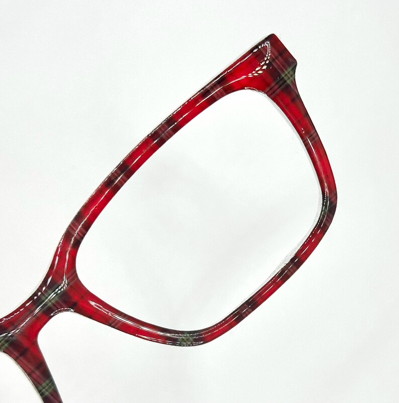 May include: Close-up of eyeglasses with a red plaid pattern. The frames are a vibrant red with black and green accents, creating a classic and stylish look. The glasses have a rectangular shape.