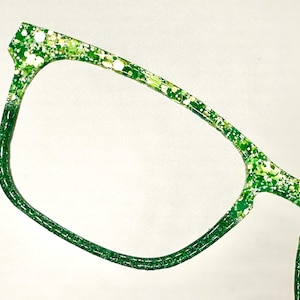 Op de afbeelding: Close-up van een bril met een groen glitterontwerp. De monturen zijn felgroen met witte en gele glitteraccenten. De bril heeft een moderne, hoekige vorm.