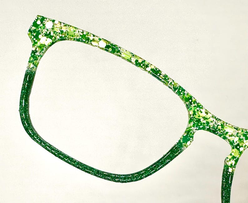 Op de afbeelding: Close-up van een bril met een groen glitterontwerp. De monturen zijn felgroen met witte en gele glitteraccenten. De bril heeft een moderne, hoekige vorm.