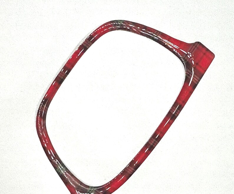 May include: A red and black plaid patterned frame, possibly for eyeglasses or a similar accessory, is set against a white background. The frame has a glossy finish and a rectangular shape with rounded corners.