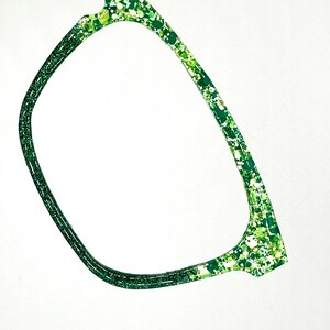 Op de afbeelding: Detail van een bril. De monturen zijn heldergroen met een gespikkeld patroon van lichtere groentinten en wit. Het ontwerp is modern en opvallend, tegen een strakke witte achtergrond.