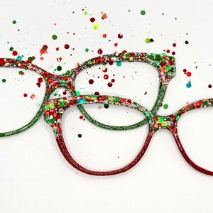 Puede incluir: Tres pares de gafas con monturas rojas, verdes y blancas brillantes. Las monturas están decoradas con purpurina de colores y salpicadas con purpurina a juego. Las gafas están sobre un fondo blanco.