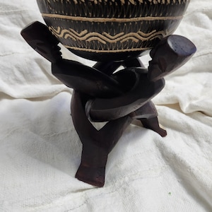 May include: A dark brown wooden bowl with carved geometric patterns in a light tan color, supported by a base featuring three interconnected human figures carved from dark wood, with each figure positioned to hold the bowl from underneath.