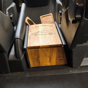 Peut inclure: Une boîte en bois avec un couvercle, conçue pour s'adapter à la console centrale d'une voiture. Le couvercle est gravé d'un motif de van avec le mot "CALIFORNIA" au-dessus.