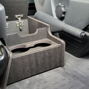 May include: A dark brown wooden cup holder with two cup slots, installed in a car's center console. The cup holder has a hinged lid that opens to reveal a storage compartment.