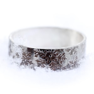 May include: A silver ring with a snowflake pattern. The ring is set against a white background with snow. The band features detailed snowflake designs, creating a winter-themed accessory. The ring's surface has a textured appearance.