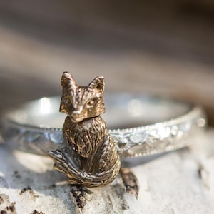 Puede incluir: Un anillo plateado con un intrincado diseño floral y una pequeña figura de zorro dorada. El zorro está en posición sentada, añadiendo un toque caprichoso a la joya.