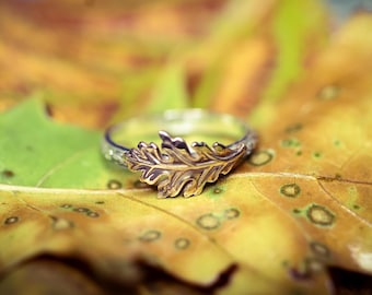 Puede incluir: Un anillo de plata con un diseño detallado de hoja de roble dorada. El anillo está sobre una hoja amarilla y verde, creando una estética inspirada en la naturaleza. La banda del anillo tiene un patrón texturizado.
